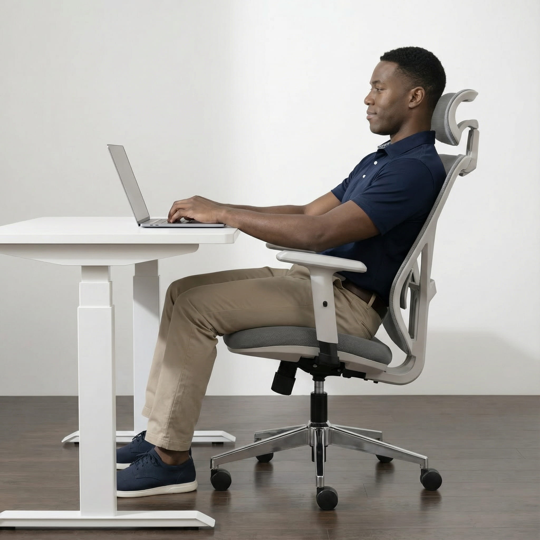 Man using a laptop on a white height adjustable desk with a ergonomic chair and mesh headrest