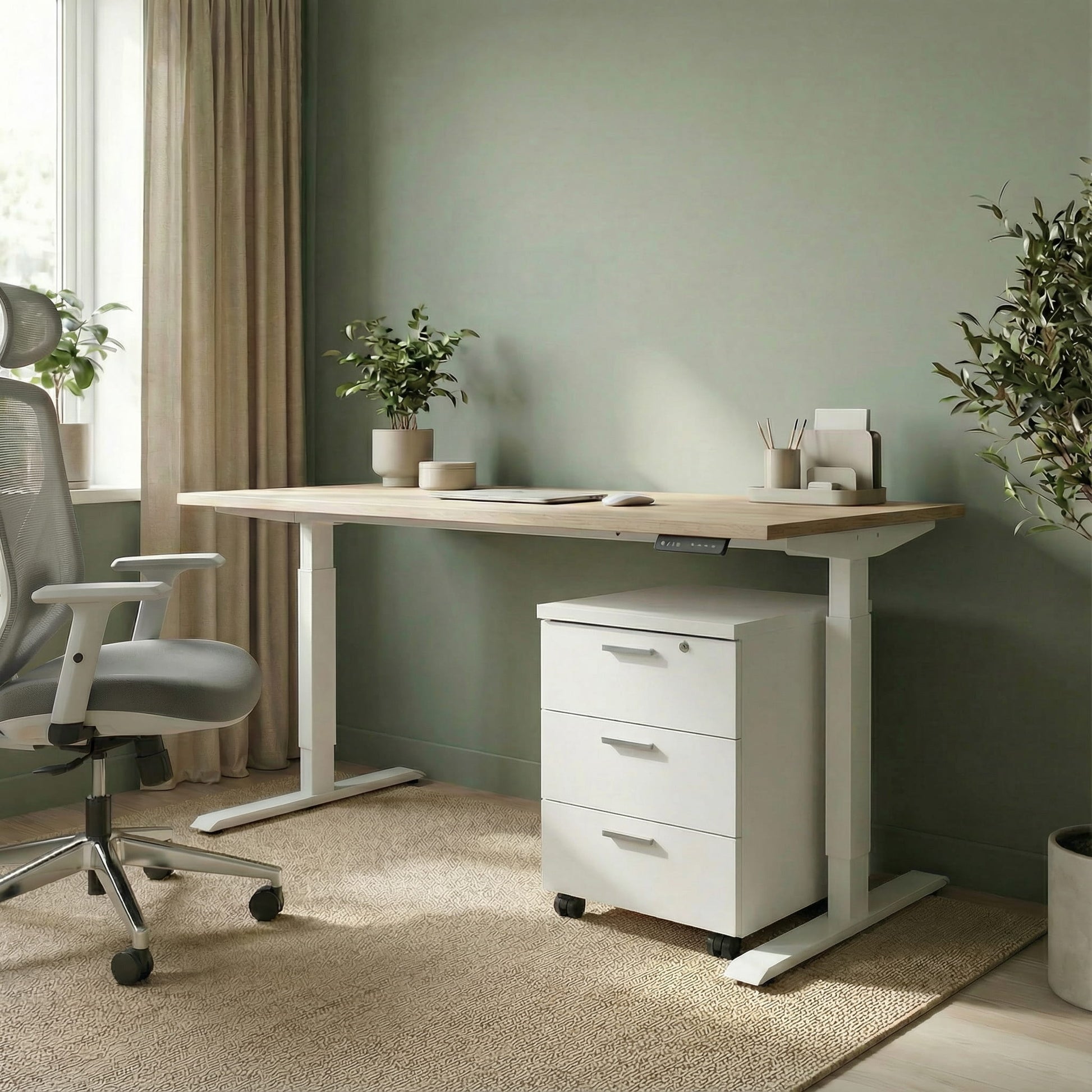 White storage with three drawers on wheels under a white framed sit stand height adjustable desk in a modern office room with a grey office chair and green background 