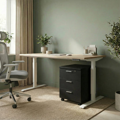 Black storage with three drawers on wheels under a white framed sit stand height adjustable desk in a modern office room with a grey ergonomic chair in the office space
