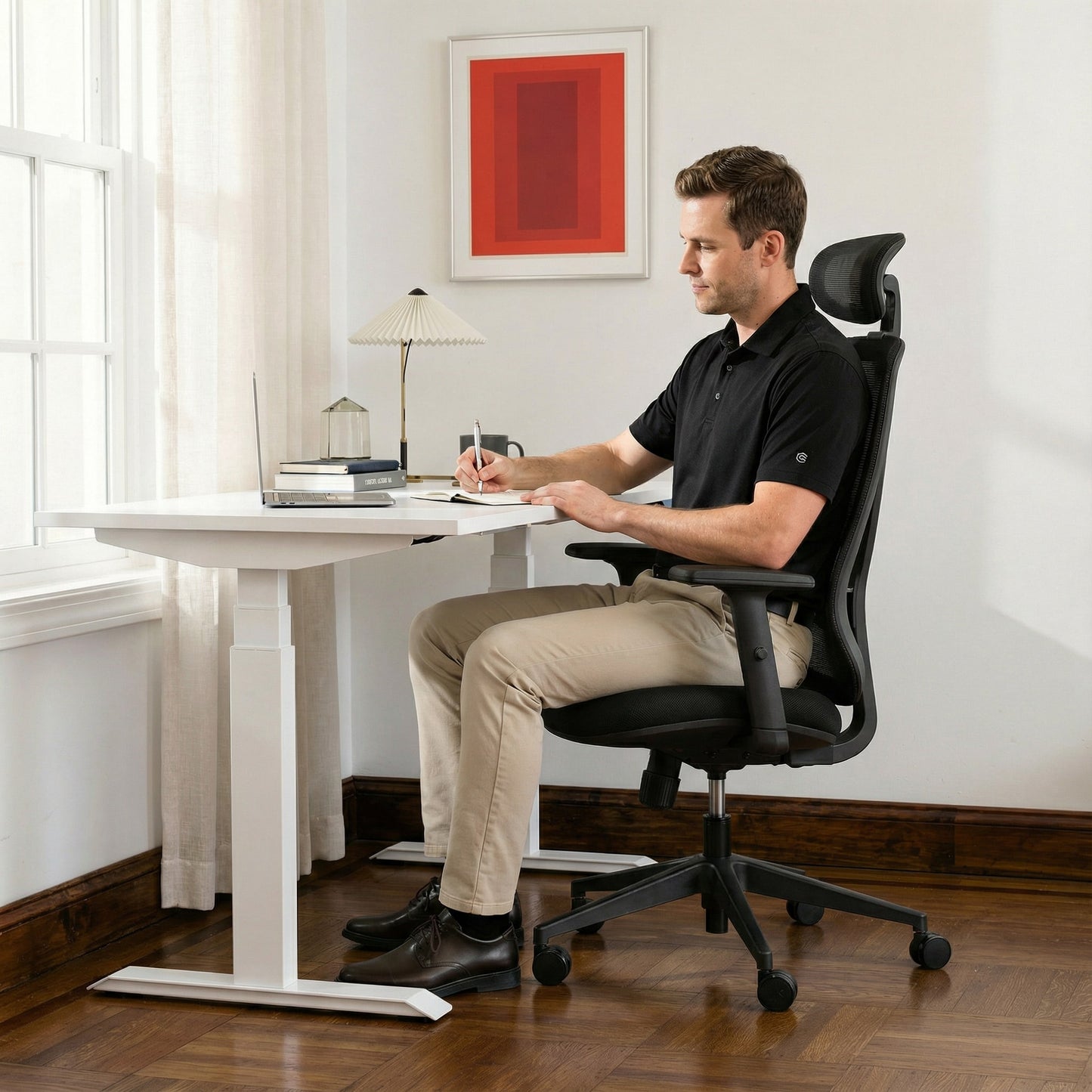 White Man sitting at a height adjustable desk in a home office with a black ergonomic office chair with mesh back and headrest.