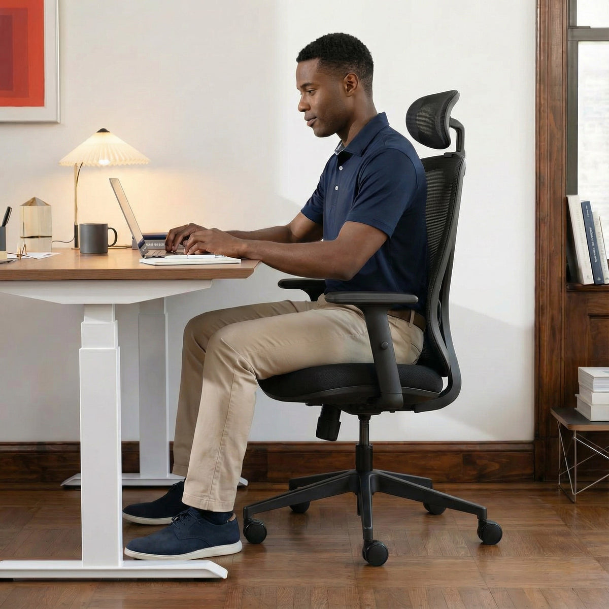 Black Man sitting at a height adjustable sit stand desk on a black ergonomic office chair using a laptop in a home office setting
